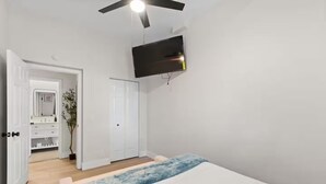 Un dormitorio con cama, ventilador de techo, televisor, armario y baño con espejo y lavamanos.