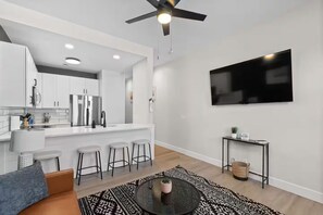 Un appartement moderne comprenant une cuisine, un coin repas et un salon. Il est équipé d’un ventilateur de plafond, d’un téléviseur à écran plat et d’une petite table d’appoint.
