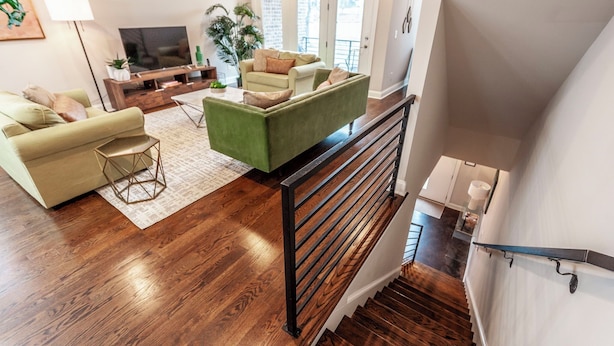 A modern living room with a green sofa, a beige sofa, and a wooden coffee table. There is a TV on a wooden stand, a potted plant, and a staircase with a black railing. - NEW! Townhome End-unit next to Geodis Park! (Nashville)