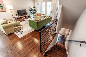 A modern living room with a green sofa, a beige sofa, and a wooden coffee table. There is a TV on a wooden stand, a potted plant, and a staircase with a black railing. - NEW! Townhome End-unit next to Geodis Park! (Nashville)