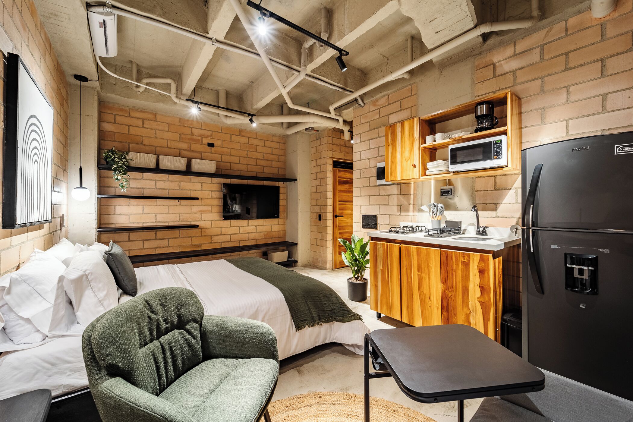 A compact studio apartment with a bed, a small seating area, a kitchenette with a microwave and refrigerator, and exposed brick walls.