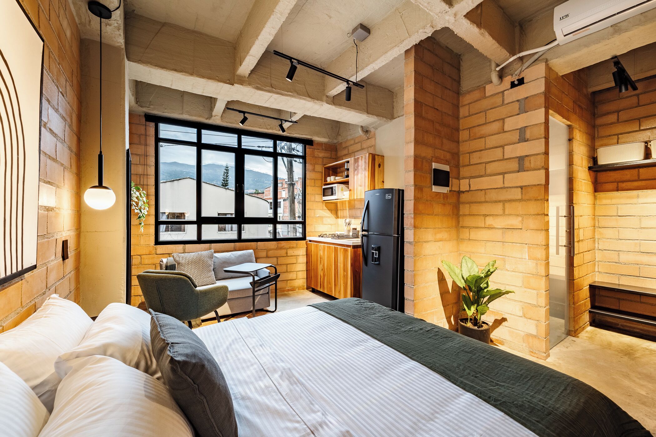 A modern room with a bed, a desk, a chair, a kitchen area, and a large window.