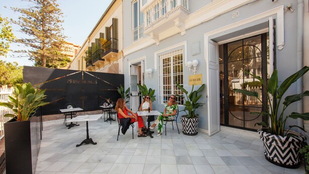 An outdoor patio area with three tables and chairs, a black sign with gold lettering, and a large potted plant. - Pries Homes - La casa azul (Málaga)