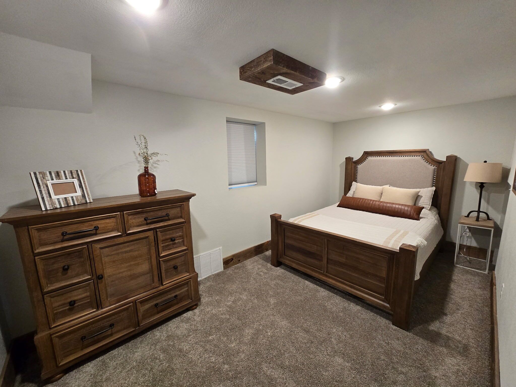 A bedroom with a wooden bed, a dresser, a nightstand, a lamp, and a window with blinds.