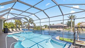 A covered outdoor swimming pool with clear blue water, surrounded by lounge chairs and a view of a river and palm trees. - Waterfront Haven of Sunshine (Cape Coral)