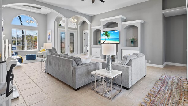 A spacious living room with a grey sofa set, a TV mounted on a white wall, and a ceiling fan. - Waterfront Haven of Sunshine (Cape Coral)