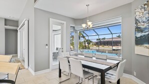 A modern dining area with a pool view, a chandelier, and a large window. - Waterfront Haven of Sunshine (Cape Coral)