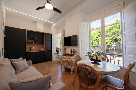 A modern living room with a sofa, dining table, and chairs. There is a kitchen area with cabinets and a television.