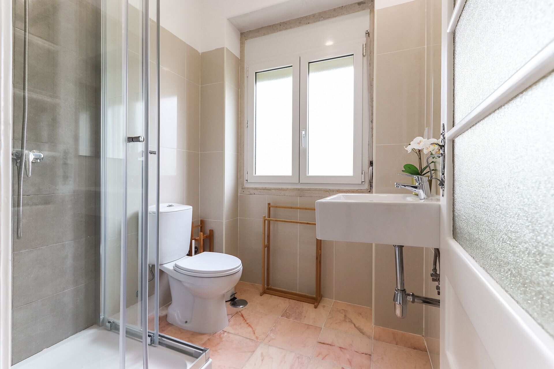 A modern bathroom with a glass shower enclosure, a toilet, a sink with a mirror, and a window above the sink.