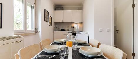 A dining area with a table set for six, white walls, and a kitchen in the background.