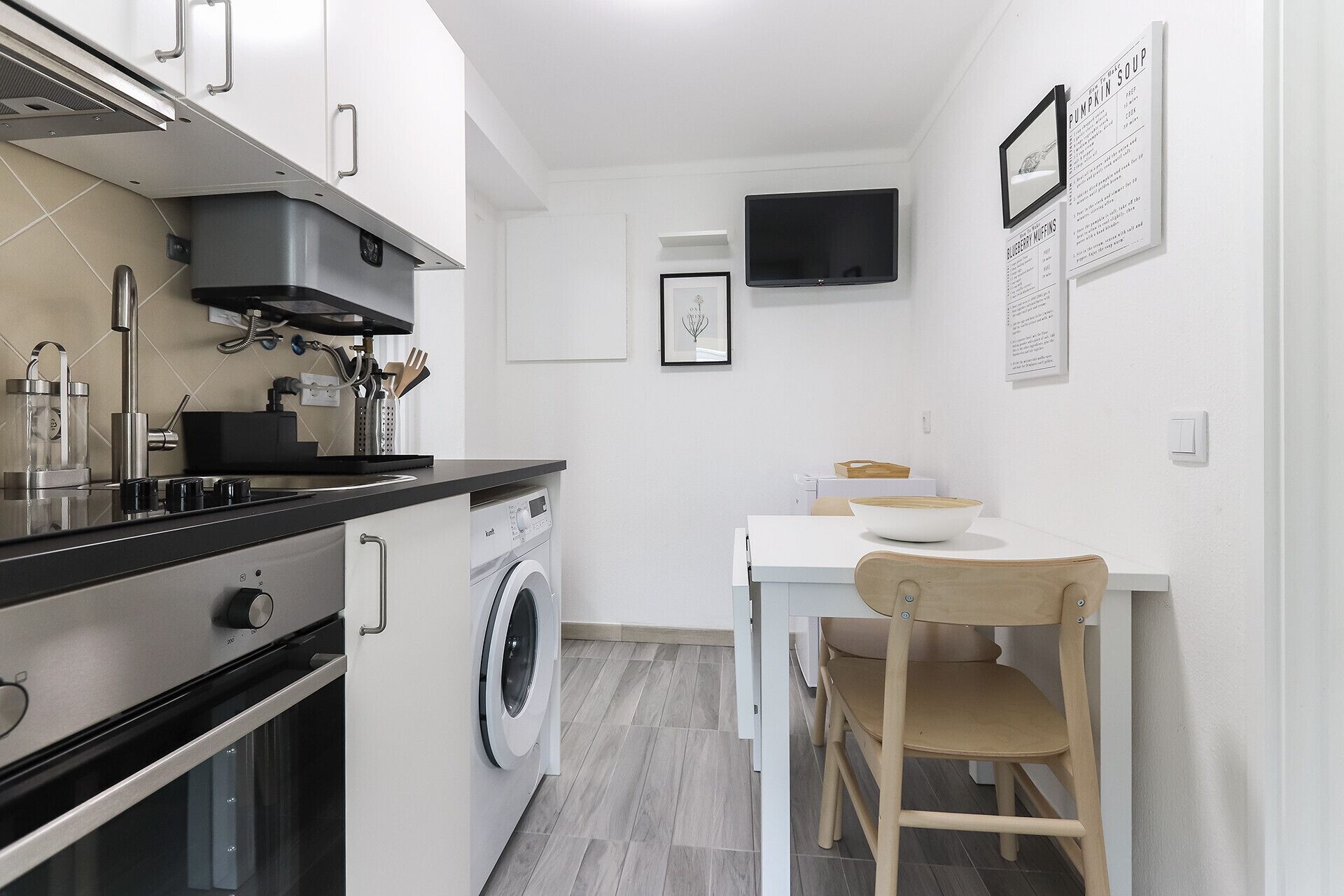 A compact kitchen with a washing machine, a dining table with a chair, and a flat-screen TV mounted on the wall.
