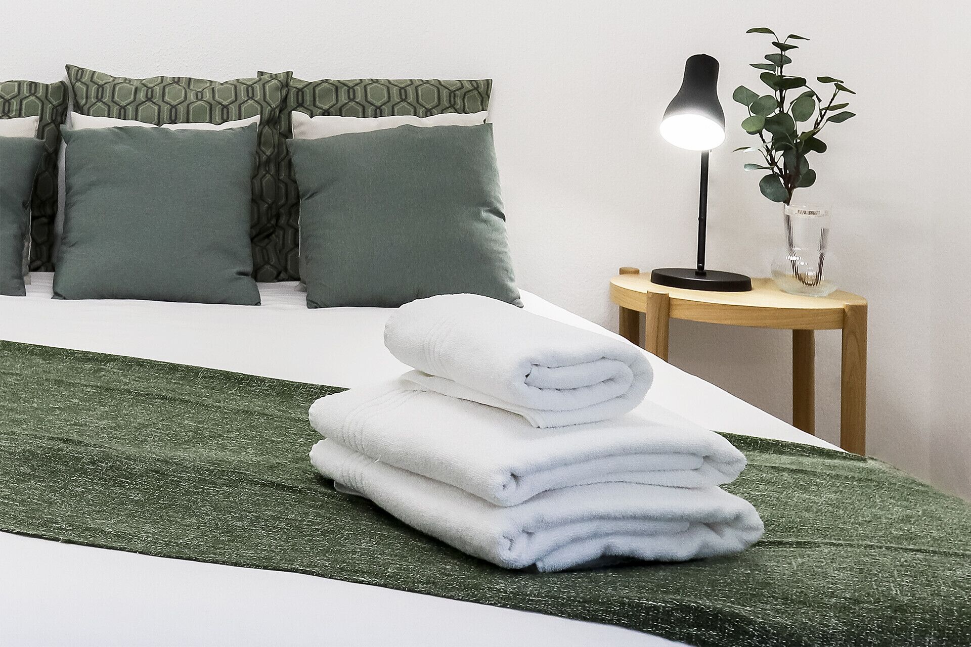 A neatly made bed with green bedding, a stack of white towels, a bedside lamp, and a small plant on a wooden nightstand.