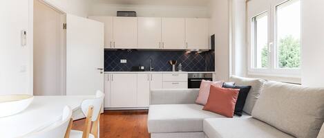 A compact kitchen with white cabinets, a black backsplash, and a built-in oven. A grey sofa with cushions is positioned near a window, and a dining table with chairs is visible.