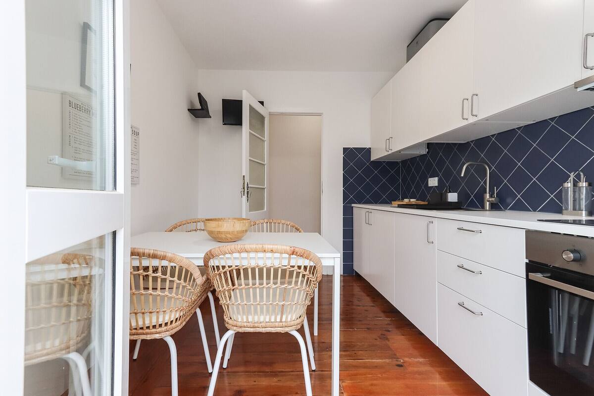 Una cucina compatta con mobili bianchi, uno schienale in piastrelle blu, un lavello in acciaio inossidabile e un forno integrato. Sono presenti sedie in vimini attorno a un piccolo tavolo con una ciotola di legno.