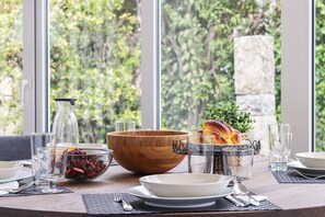 A dining table set with clear glassware, a wooden bowl, and a croissant, with a view of greenery through large windows. - Nine Horizons Belém (Lisbon)