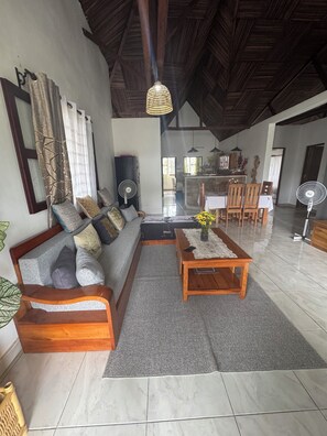 A spacious living area with a large wooden sofa, a coffee table, and a dining table. There is a kitchen in the background with a dining area. - CHARMING VILLA WITH GARDEN AND STAFF NEAR THE BEACHES; LONG-TERM RENTAL AVAILABLE. (nosy be)