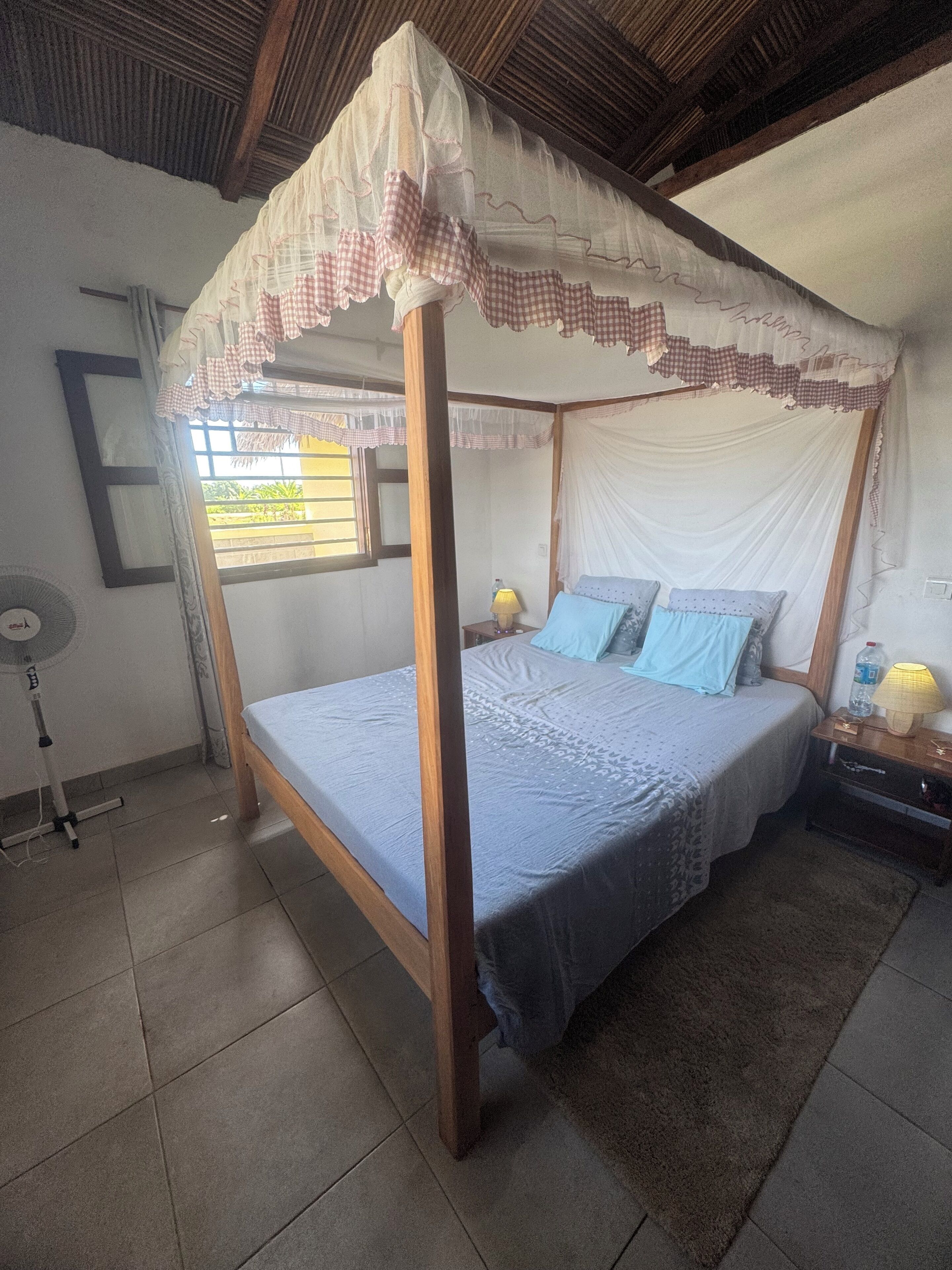 Uma cama com dossel e cortina, uma mesa de cabeceira com abajur e um ventilador em um quarto com piso em azulejos.