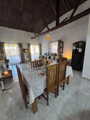 A dining area with a table set for four, wooden chairs, a ceiling fan, and a refrigerator. - CHARMING VILLA WITH GARDEN AND HOUSE STAFF IN NOSY BÉ, CLOSE TO THE BEACHES (nosy be)