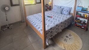 A bedroom with a four-poster bed, a standing fan, a small table with a lamp, a bookshelf, and a window with curtains. - CHARMING VILLA WITH GARDEN AND HOUSE STAFF IN NOSY BÉ, CLOSE TO THE BEACHES (nosy be)