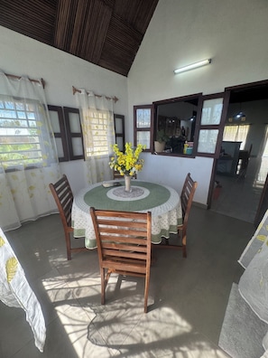 A dining area with a round table, four wooden chairs, a vase with yellow flowers, and a view of a kitchen through a window. - CHARMING VILLA WITH GARDEN AND STAFF NEAR THE BEACHES; LONG-TERM RENTAL AVAILABLE. (nosy be)