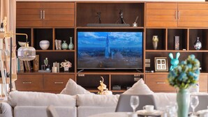 Un soggiorno moderno con un'ampia televisione a schermo piatto, un divano, un tavolo da pranzo per due persone e un mobile in legno con oggetti decorativi.