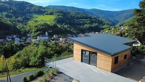 A modern house with a large glass window and a metal roof, surrounded by lush greenery and a village in the background. - Luxury Chalet with Sauna and Hot Tub (Fréland)