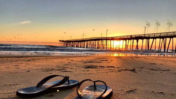 Escena de playa con un muelle al fondo y un par de chanclas en primer plano.