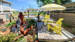 Una terraza de madera con sillas Adirondack amarillas, un pequeño jardín con una escultura de pez, y una pérgola con un paraguas amarillo.