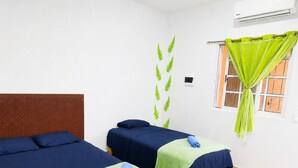 Een kamer met twee bedden, elk met een blauwe spreideken en een groene handdoek. Er is een raam met een groene gordijn, en een luchtverkoeler aan de muur bevestigd.
