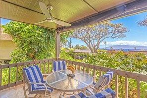 Ein überdachter Außenbereich mit Tisch und Stühlen, umgeben von einer Brüstung, mit Blick auf einen üppigen Garten und das Meer.