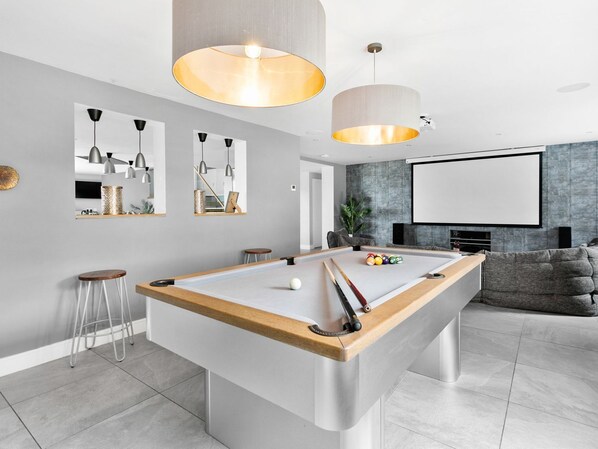 A modern living room with a pool table, bar stools, and a large screen for entertainment.