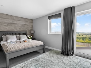 A modern bedroom with a large bed, a view of the countryside, and a window with curtains. - Huxham View and Annex (Exeter)