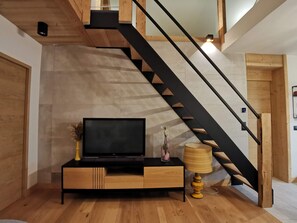 A modern living room with a wooden floor, a black TV stand, a lamp, and a staircase with wooden steps and black railings. - Assy's Cocoon (Passy)