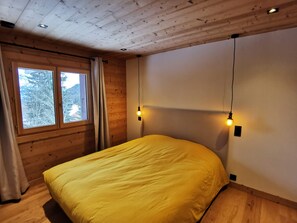 A room with a bed, wooden walls, a window with curtains, and two wall-mounted lights. - Assy's Cocoon (Passy)