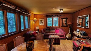 Un salon chaleureux avec une table à manger en bois, des sièges confortables et de grandes fenêtres donnant sur l’extérieur.