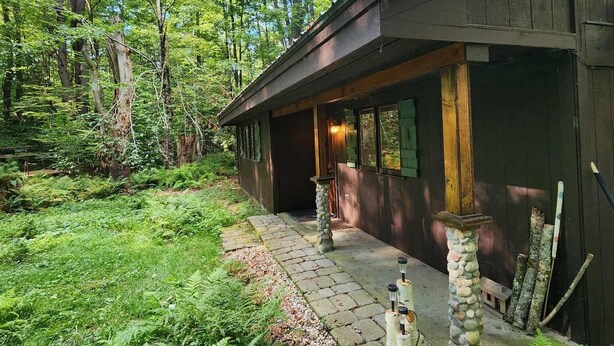 Un chalet rustique avec un sentier de pierres menant à l’entrée, entouré d’une végétation luxuriante et de grands arbres.