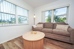 A living room with a brown sofa, a round wooden coffee table, and large windows with blinds.