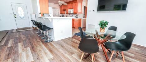A modern kitchen with wooden cabinets, a microwave, and a ceiling fan. There is a dining area with a glass table and black chairs.