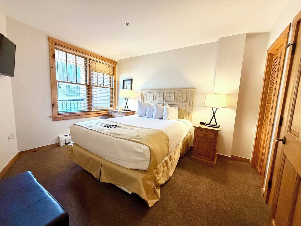 A bedroom with a large bed, a bedside table, a lamp, a window with blinds, and a door.