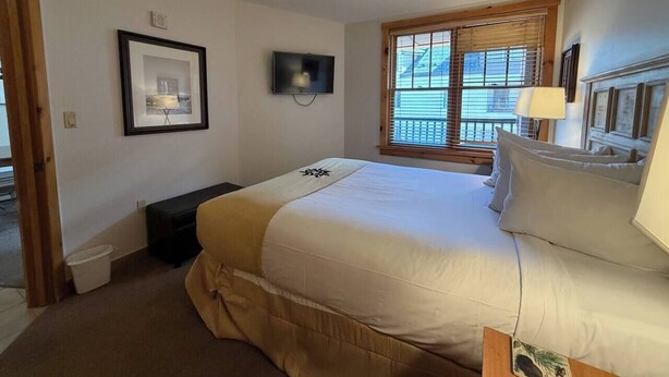 A bedroom with a large bed, a television mounted on the wall, a window with blinds, and a framed picture on the wall. - Keystone Mountain Escape (Keystone)