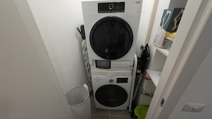 A compact laundry room with a front-loading washer and dryer stacked on top of each other, a small trash bin, and a shelf with a box and other items. - A Room with private bathroom; Sweet Home Fleming (Roma)
