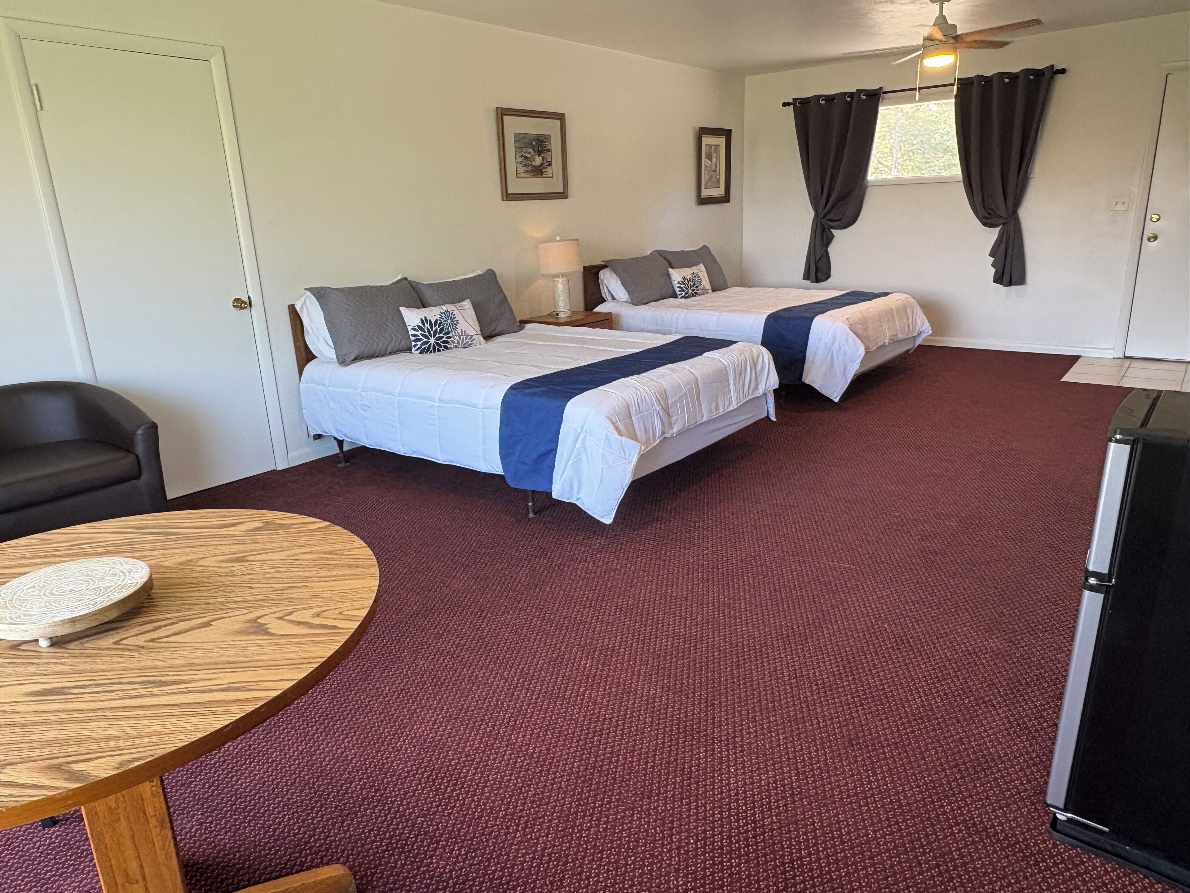 A hotel room with two beds, a desk, a chair, a table, and a refrigerator.