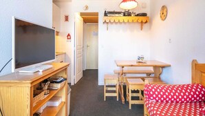 Ein gemütlicher Raum mit Fernseher auf einem Holzboden, einem kleinen Tisch mit Bänken, einem roten Sofa mit Punkten und einem Regal mit einer Lampe.