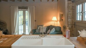 Un baño rústico con un lavamanos grande de color blanco, vigas de techo de madera, una pared de piedra y una vista de un sofá verde al fondo.