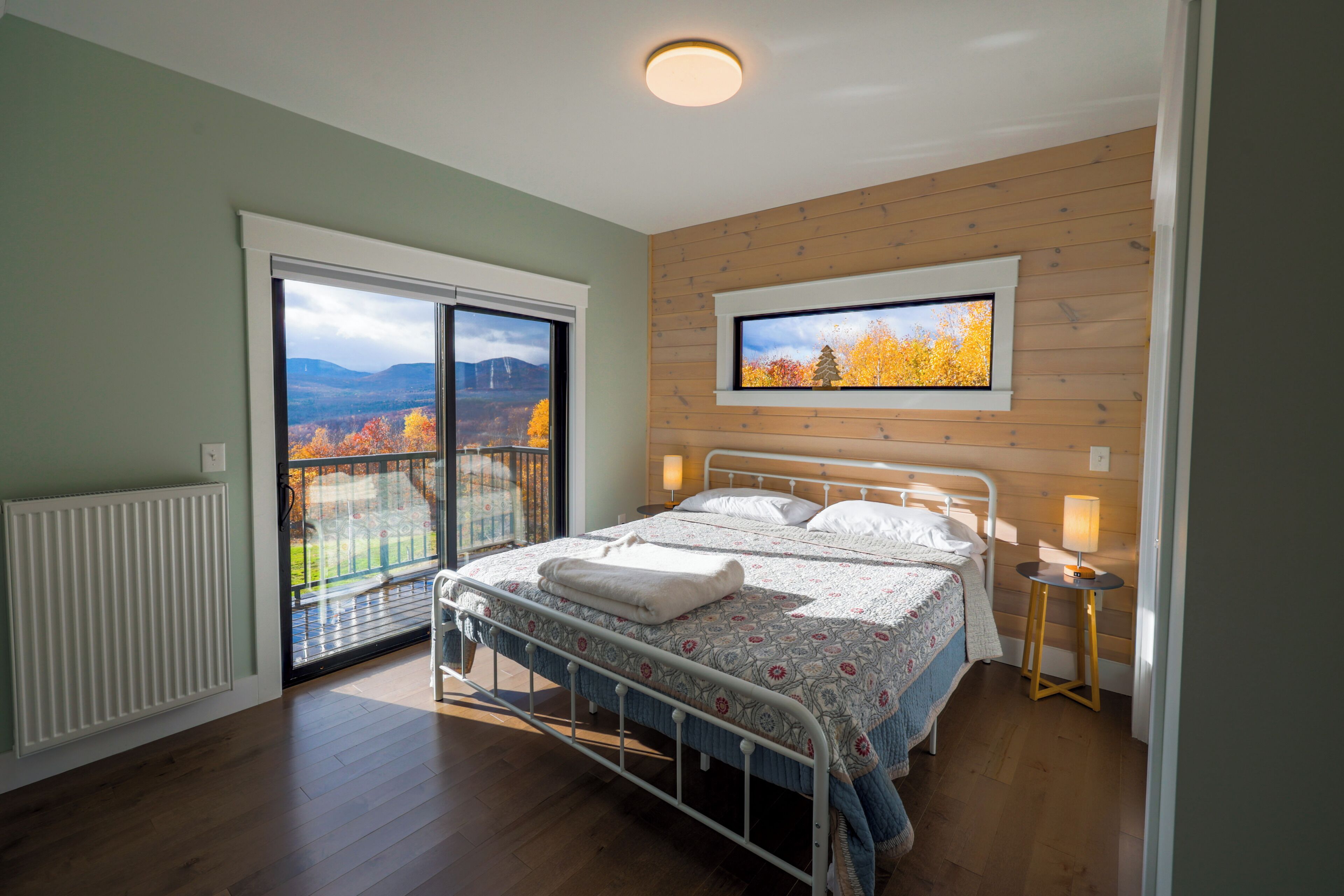 A bedroom with a bed, a TV mounted on the wall, a window with a view of autumn trees, and a balcony with a railing.