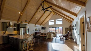 A spacious kitchen with wooden beams, a dining area, and a living room with a stone wall. - Mountain Escape - Views • Firepit • Near Sugarloaf (Kingfield)