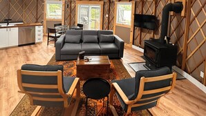 A cozy, open-plan living space with a high ceiling, wooden beams, a sofa, chairs, a coffee table, a fireplace, and a kitchen area. - Yurtopia on the Lake (Lanark Highlands)