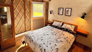 A bedroom with a sloped ceiling, wooden beams, a bed with a tree-patterned bedspread, a window, and framed pictures on the wall. - Yurtopia on the Lake (Lanark Highlands)