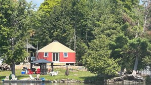 A lakeside cabin with a red exterior, surrounded by trees and a dock with chairs. - Yurtopia on the Lake (Lanark Highlands)