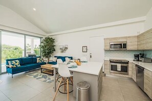 A modern kitchen with a central island, a dining area with a blue sofa, and a large window providing a view of greenery. - Azure Aruba-3BR Resort Style Villa by Eagle Beach (Oranjestad)